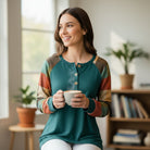 Woman wearing HIGHLAND RETREAT TOP with colorful sleeves while enjoying a cup of coffee indoors.