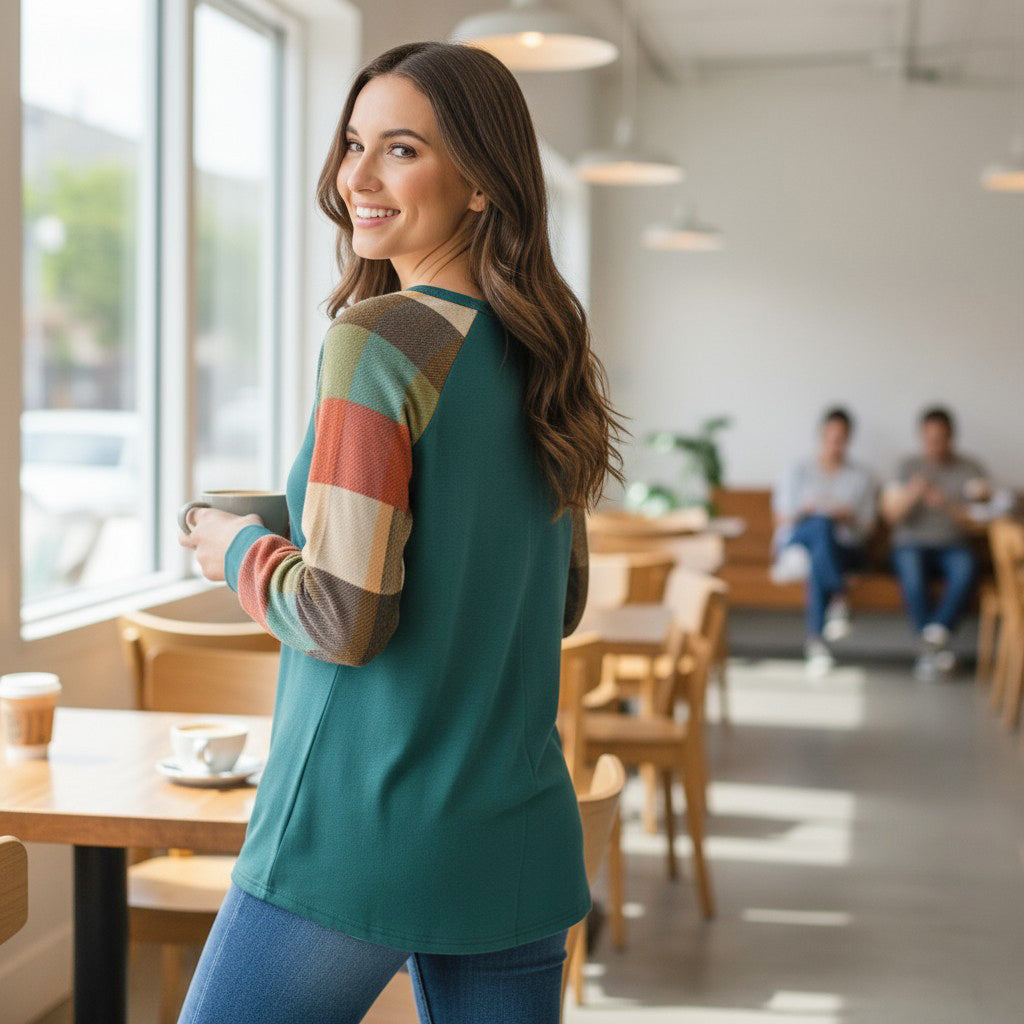 Woman in a cozy cafe wearing the HIGHLAND RETREAT TOP with plaid sleeves, enjoying a cup of coffee.